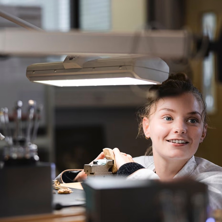 Young watchmaker smiling while working at a desk with watchmaking tools and equipment.