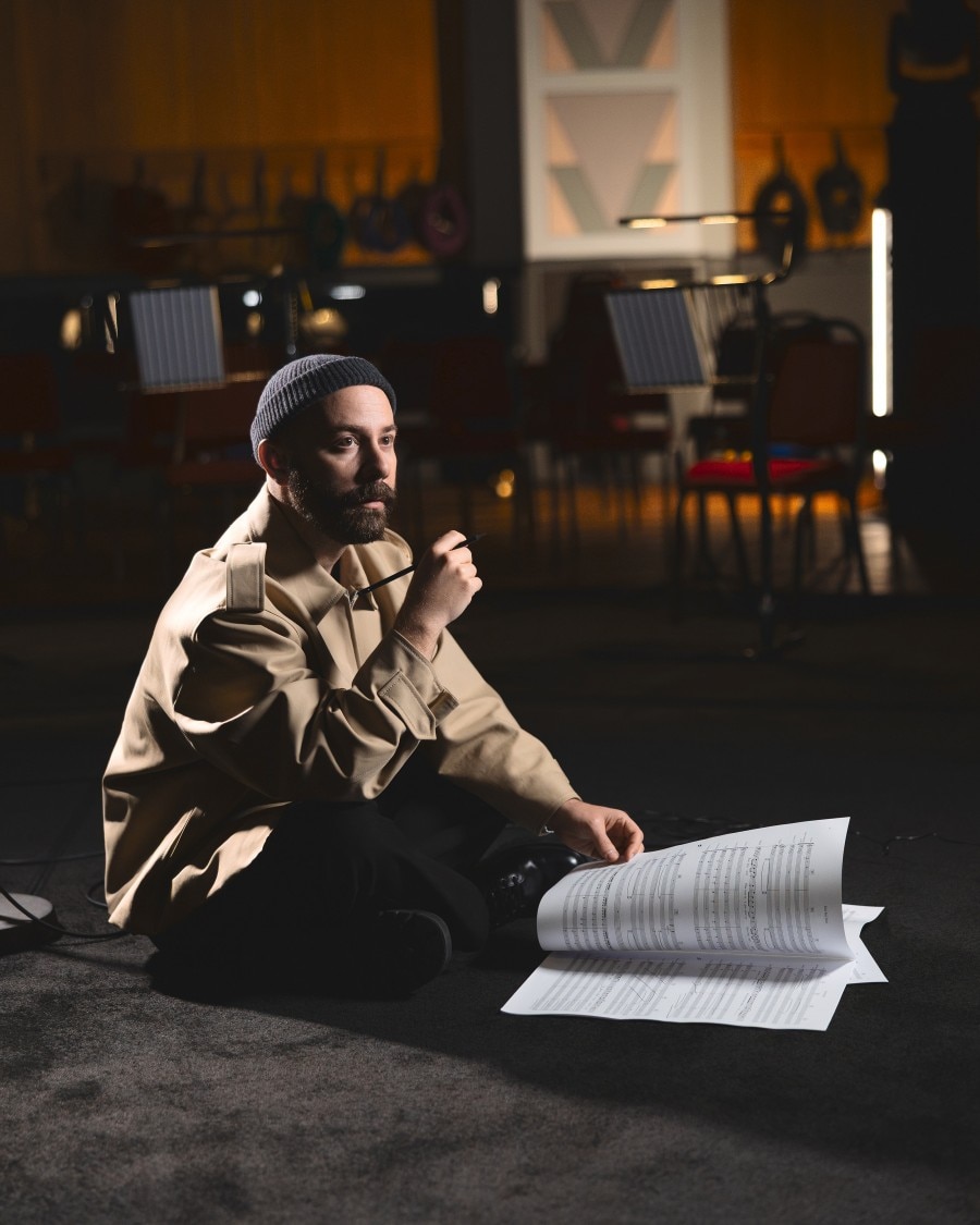 Woodkid sitting on the floor with sheet music, holding a pencil in a rehearsal space.
