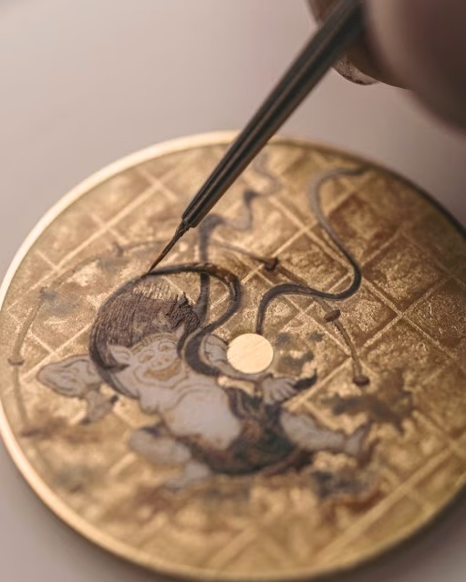 Artisan using tweezers to place decorative elements on a watch dial featuring a traditional Asian-style painting.