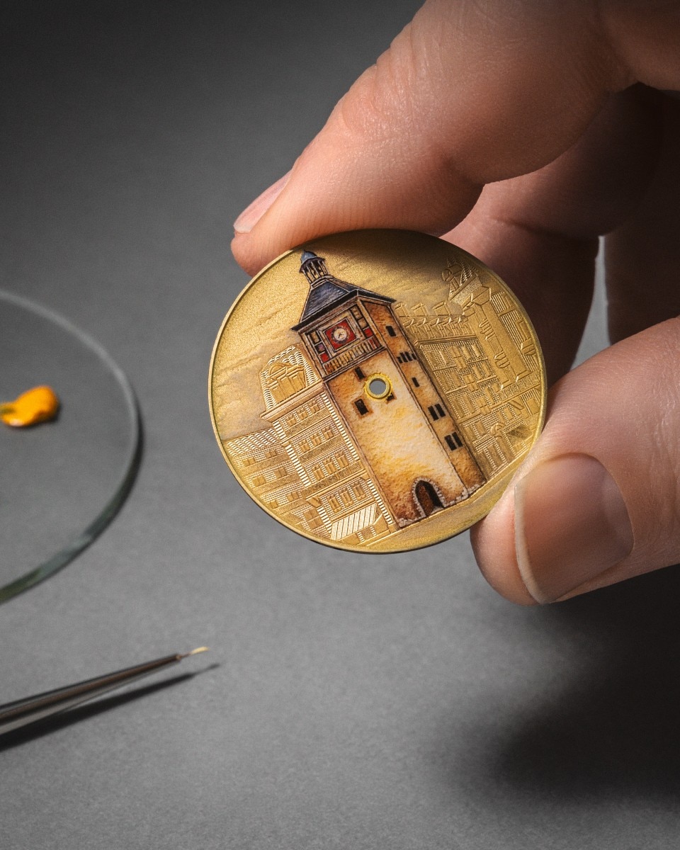 Artisan holding a painted watch dial featuring a yellow background and a clock tower illustration.