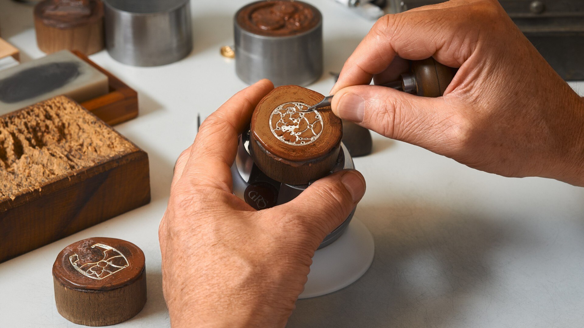 Hands engraving a decorative motif into a small round metal surface