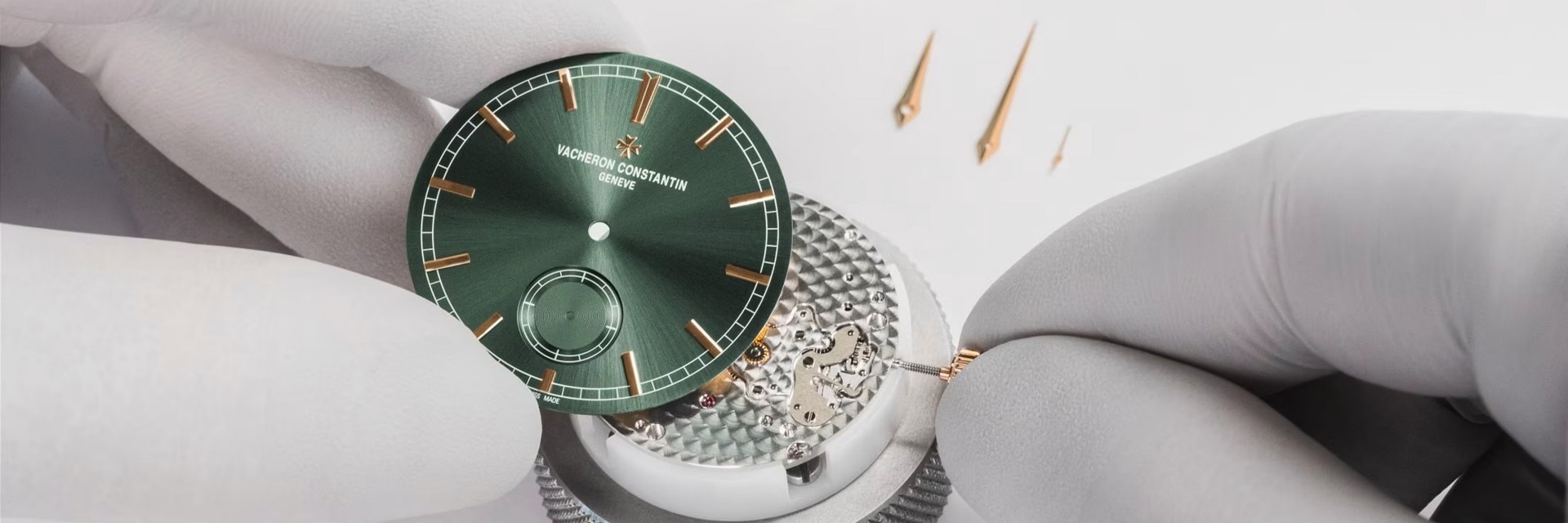 Two hands in white gloves are setting up a watch with a green dial with gold details