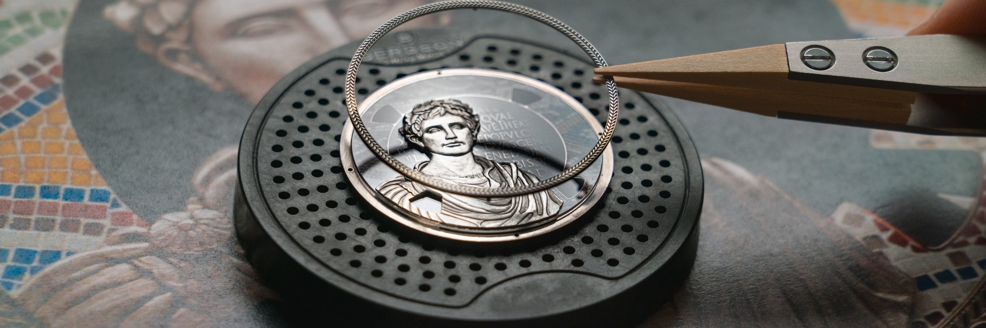 Tweezers holding a decorative ring above a detailed watch dial featuring a Roman bust.