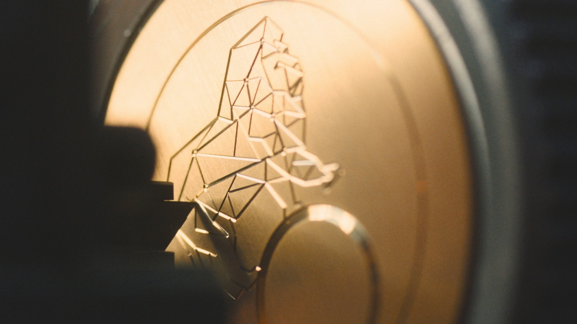 Close-up of a gold watch component featuring a geometric lion relief engraving, partially in shadow.