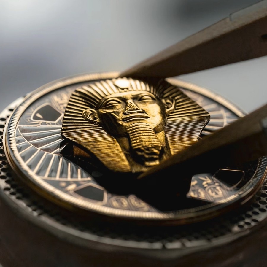 Close-up of tweezers placing a golden pharaoh head sculpture onto a watch dial.