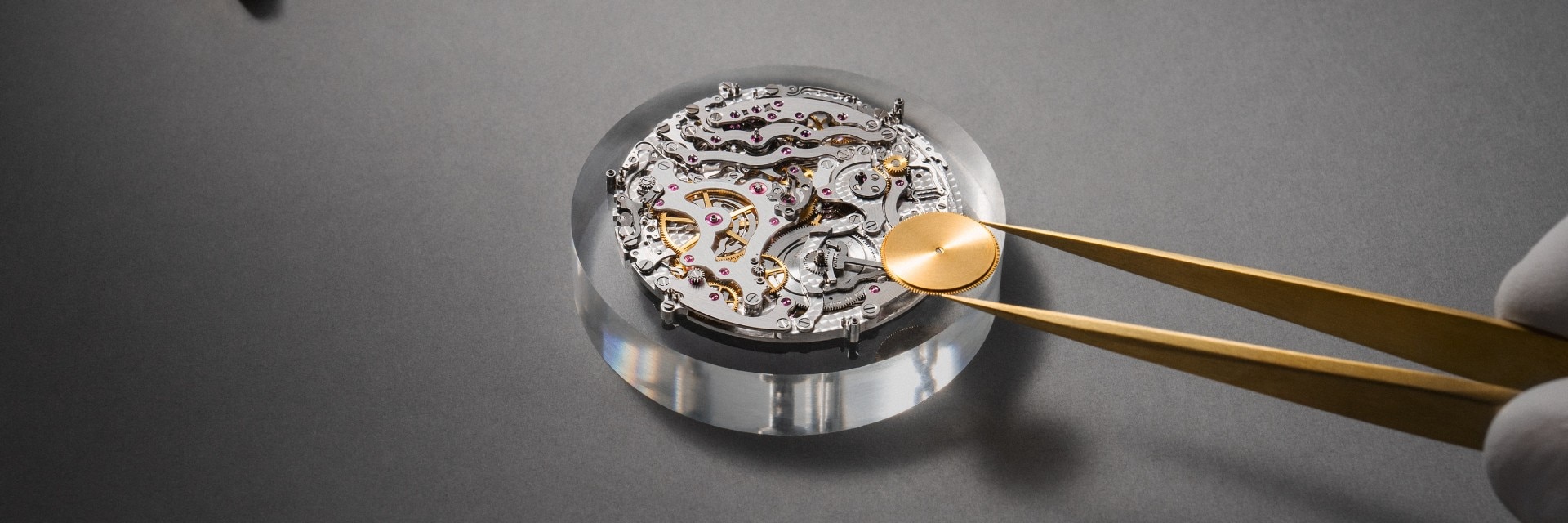 Tweezers holding a gold piece over an intricate watch movement.