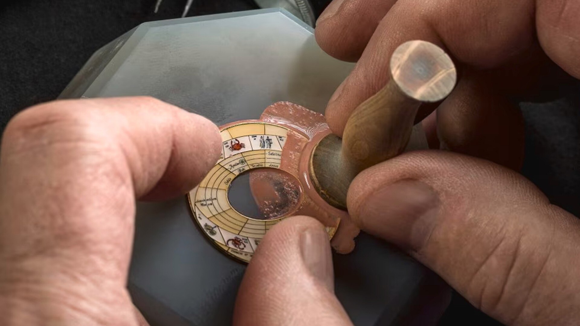 Craftsman's hands engraving and painting intricate zodiac symbols onto a small circular watch component using a precision tool.
