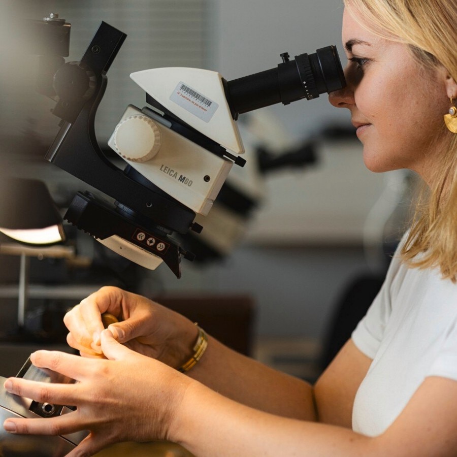 Woman examining a watch component under a microscope in a watchmaking workshop.