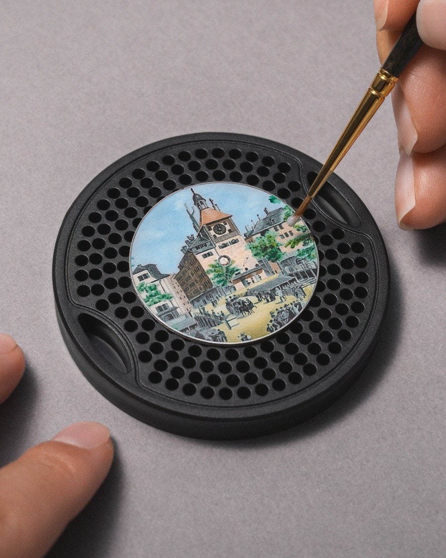 Artist hand-painting an enamel watch dial with a historic city scene.