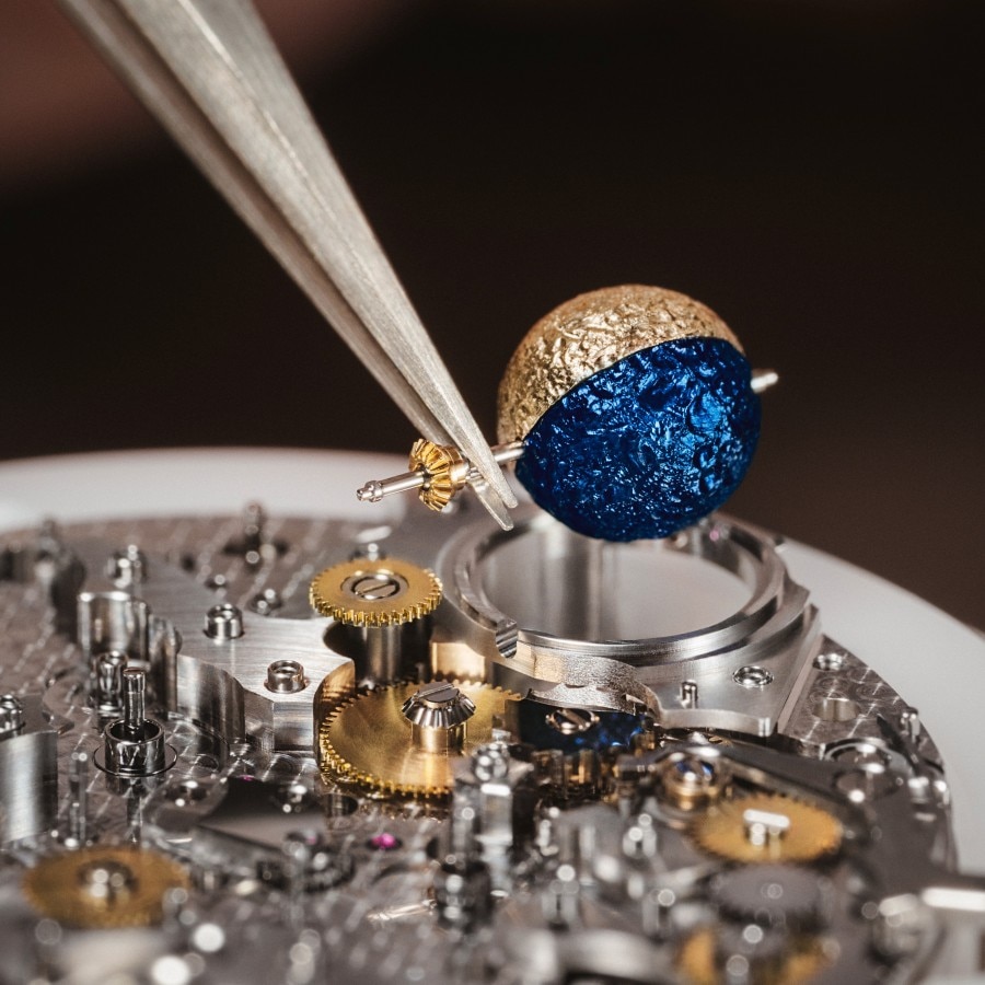 Watchmaker assembling a round, gold and blue element onto a watch movement with tweezers.