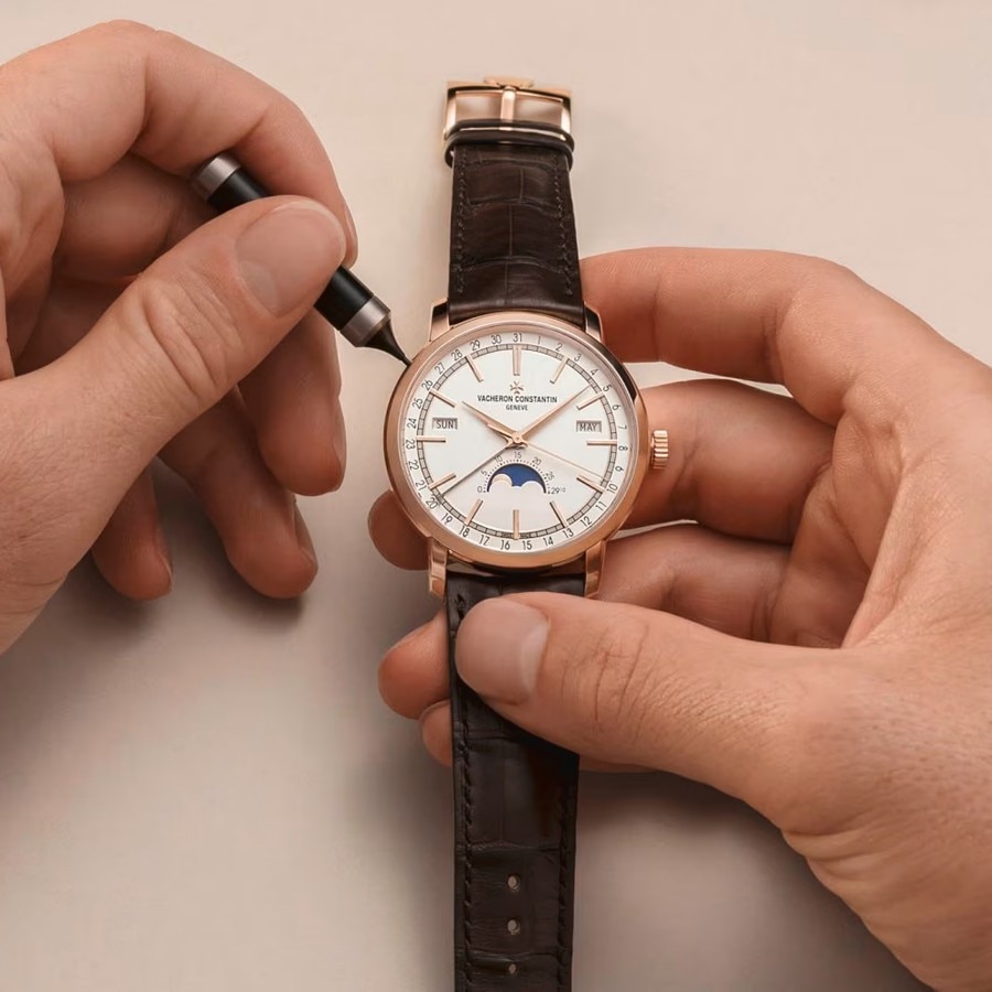 Two human hands are setting up a watch with a leather strap and a white dial with gold hands and frame