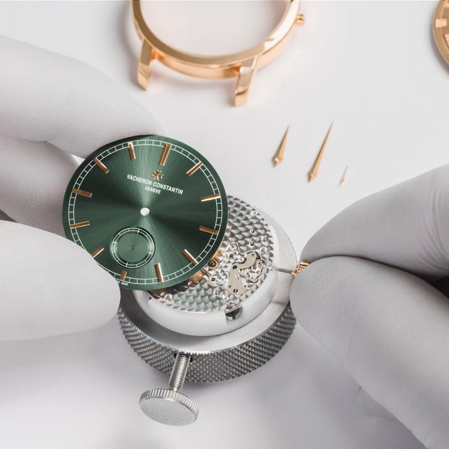 Two hands in white gloves are setting up a watch with a green dial with gold details