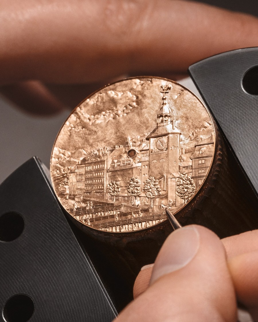 Hand holding a golden engraved watch dial with detailed architectural elements.