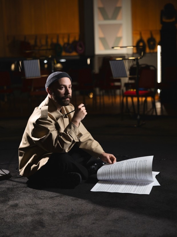 Woodkid sitting on the floor with sheet music, holding a pencil in a rehearsal space.