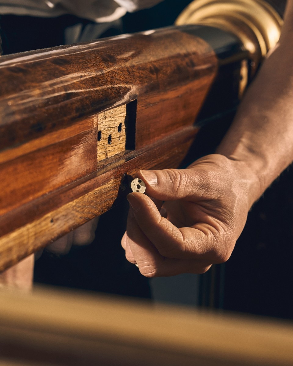 Hand holding a small metal piece next to a wooden lock plate on antique furniture.
