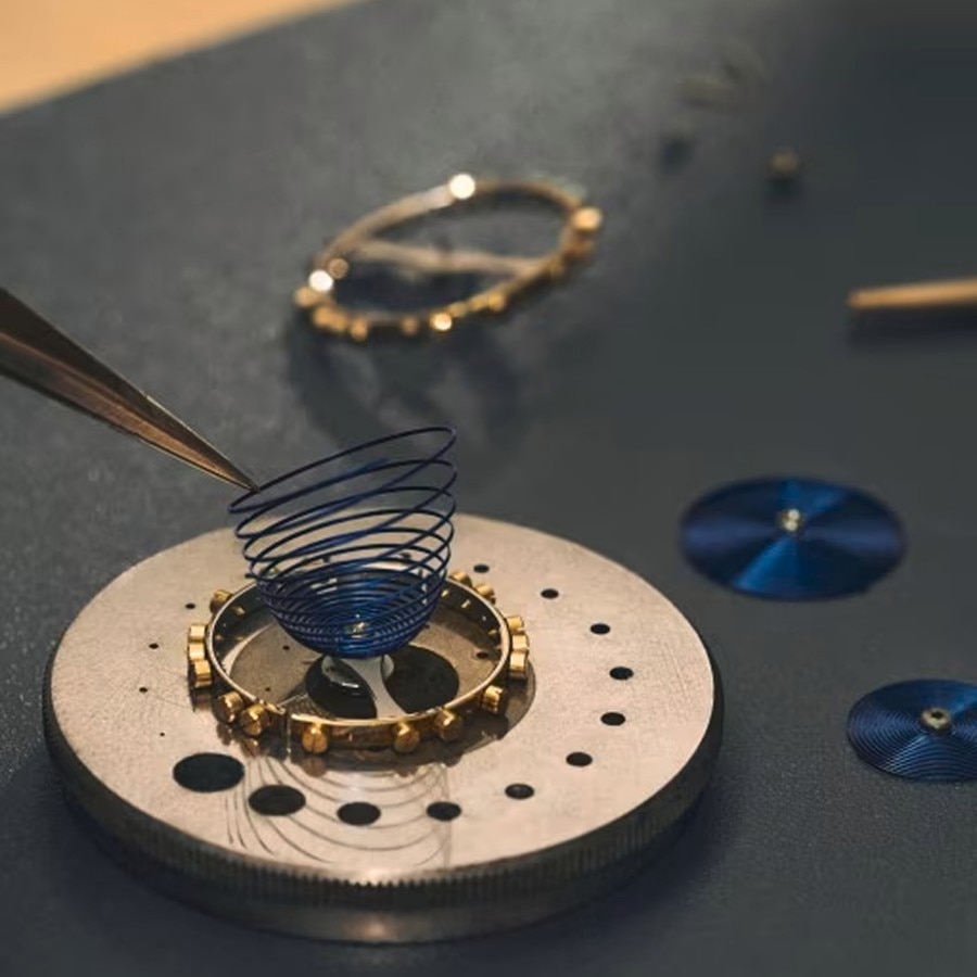 Watchmaking components on a workbench, with tweezers holding a delicate hairspring above a gear assembly.