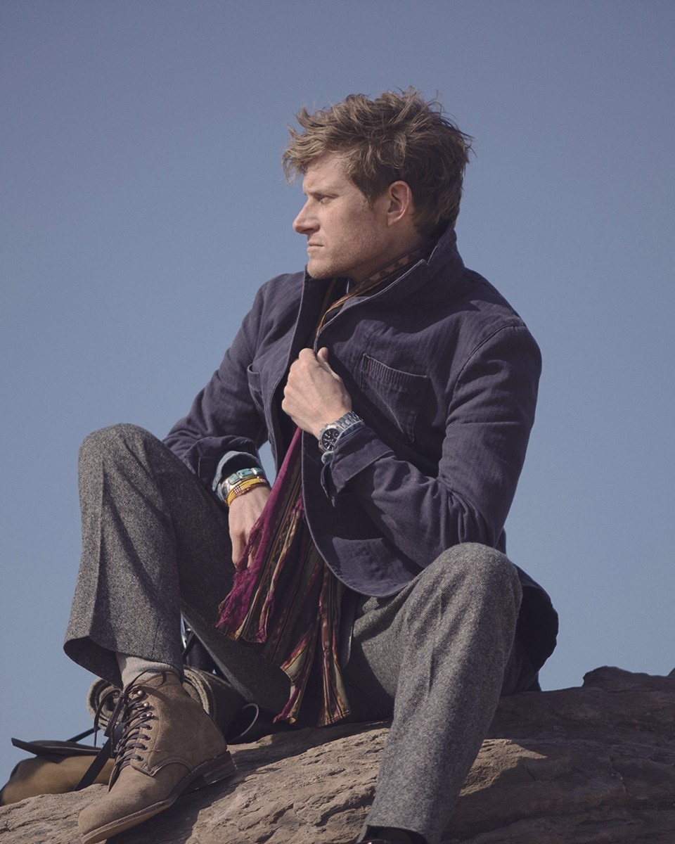 Man in outdoor wear sitting on a rock, looking to the side while holding a watch.