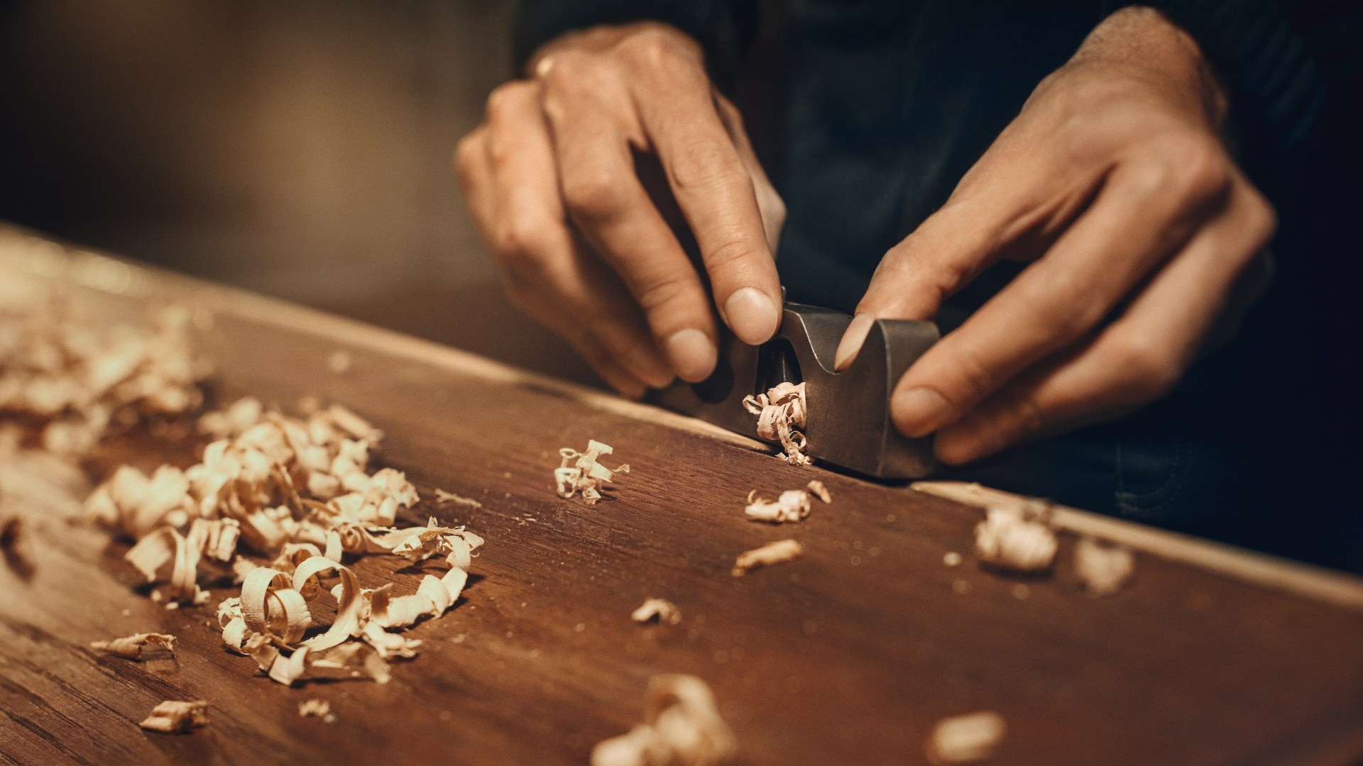 Close-up of hands carving wood shavings on a wooden surface using a chisel.