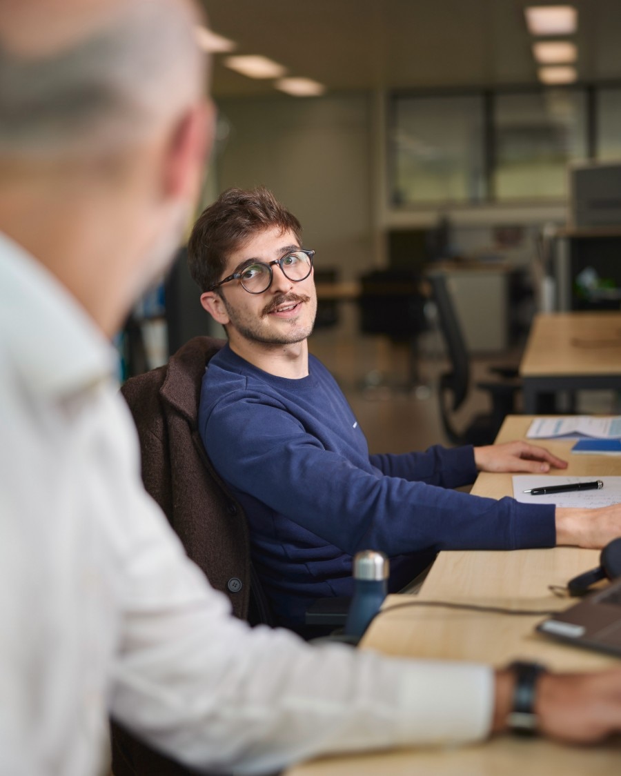 Two men having a conversation in an office, one seated at a desk and smiling.