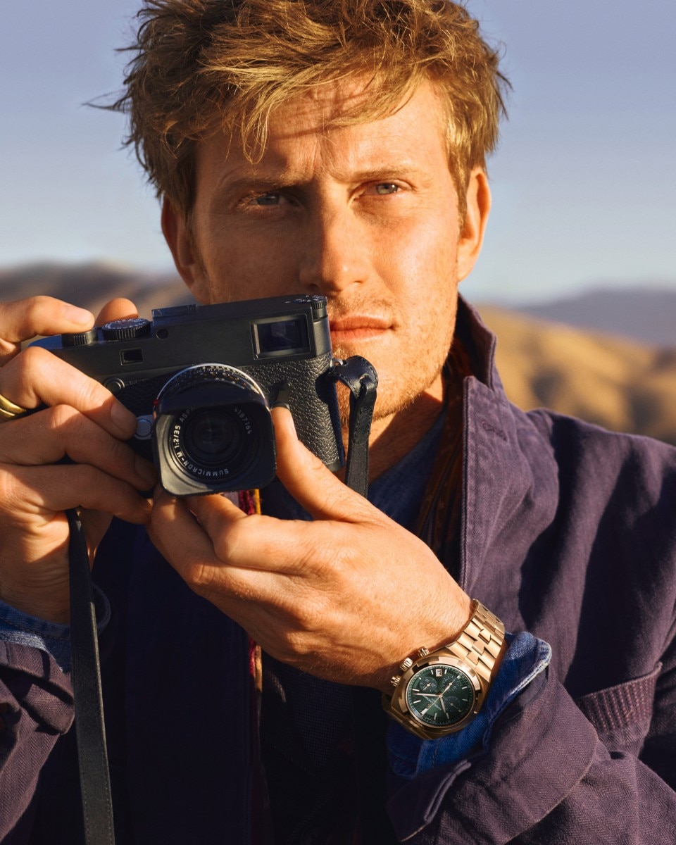 Man holding a camera in a desert landscape with mountains in the background.