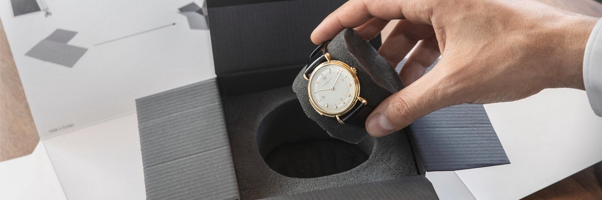 Hand placing a gold watch into a foam insert inside a presentation box.