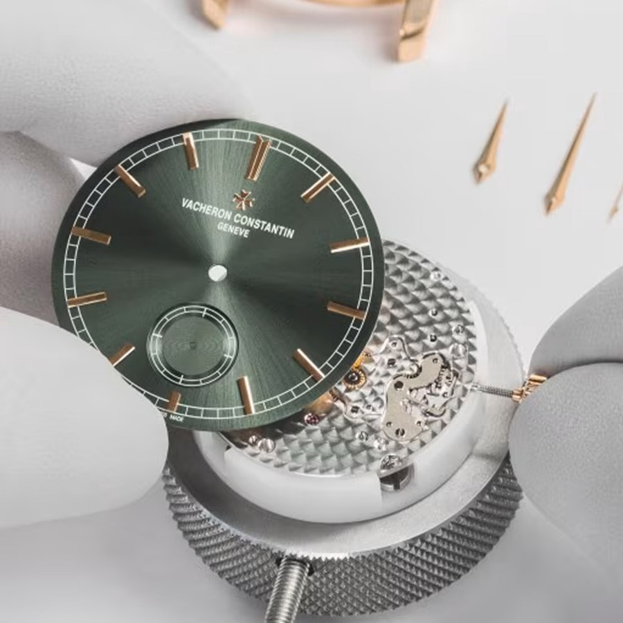 Watchmaker assembling a movement while holding a green dial with gloved hands.