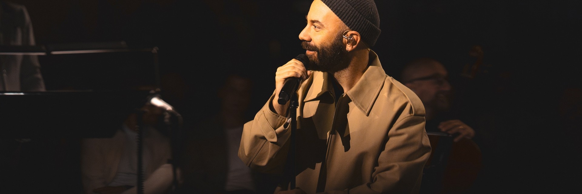 Woodkid holding a microphone and wearing a beanie and coat, performing on stage.