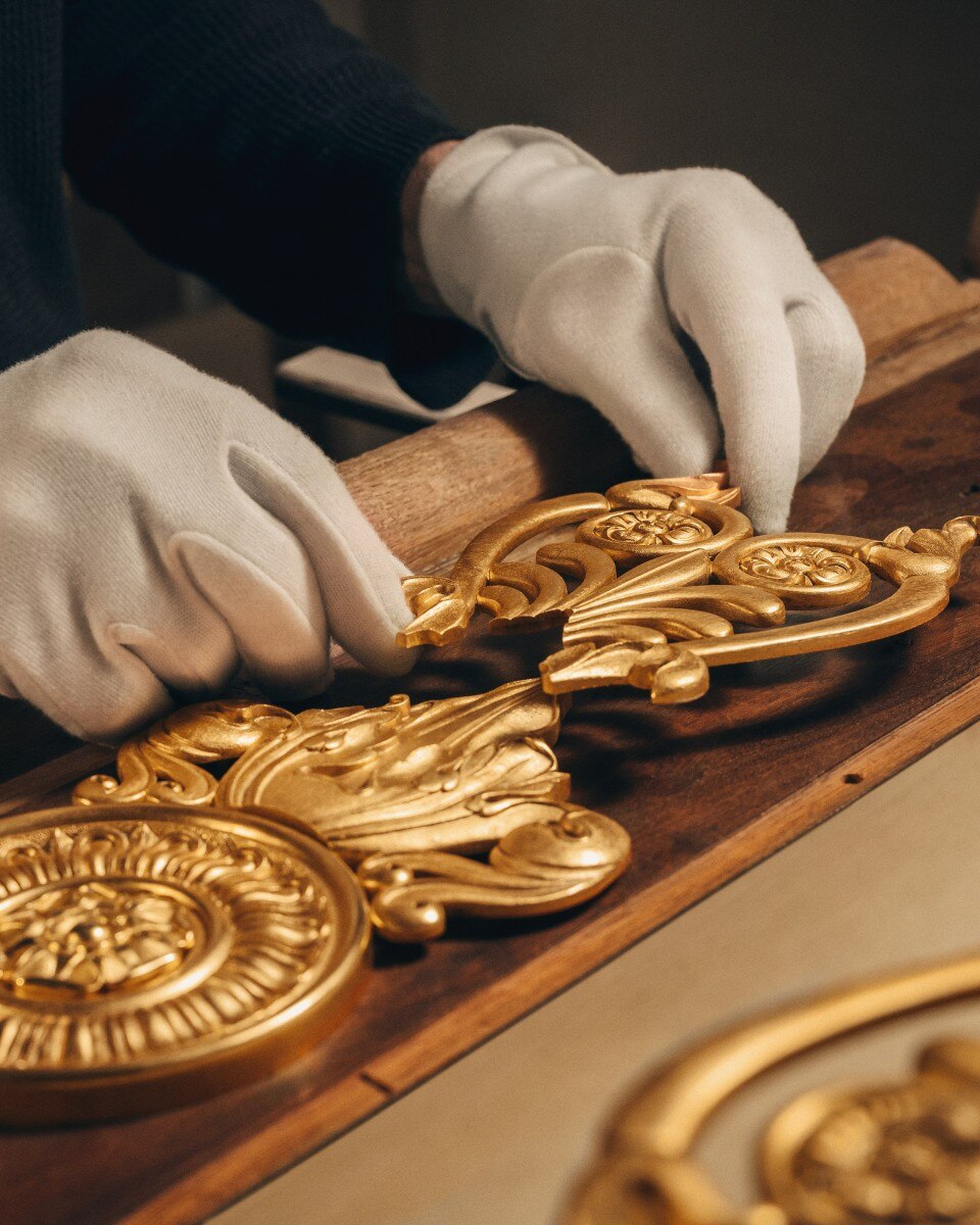 Close-up of a gloved hands restoring or assembling intricate golden clock components on a wooden surface.