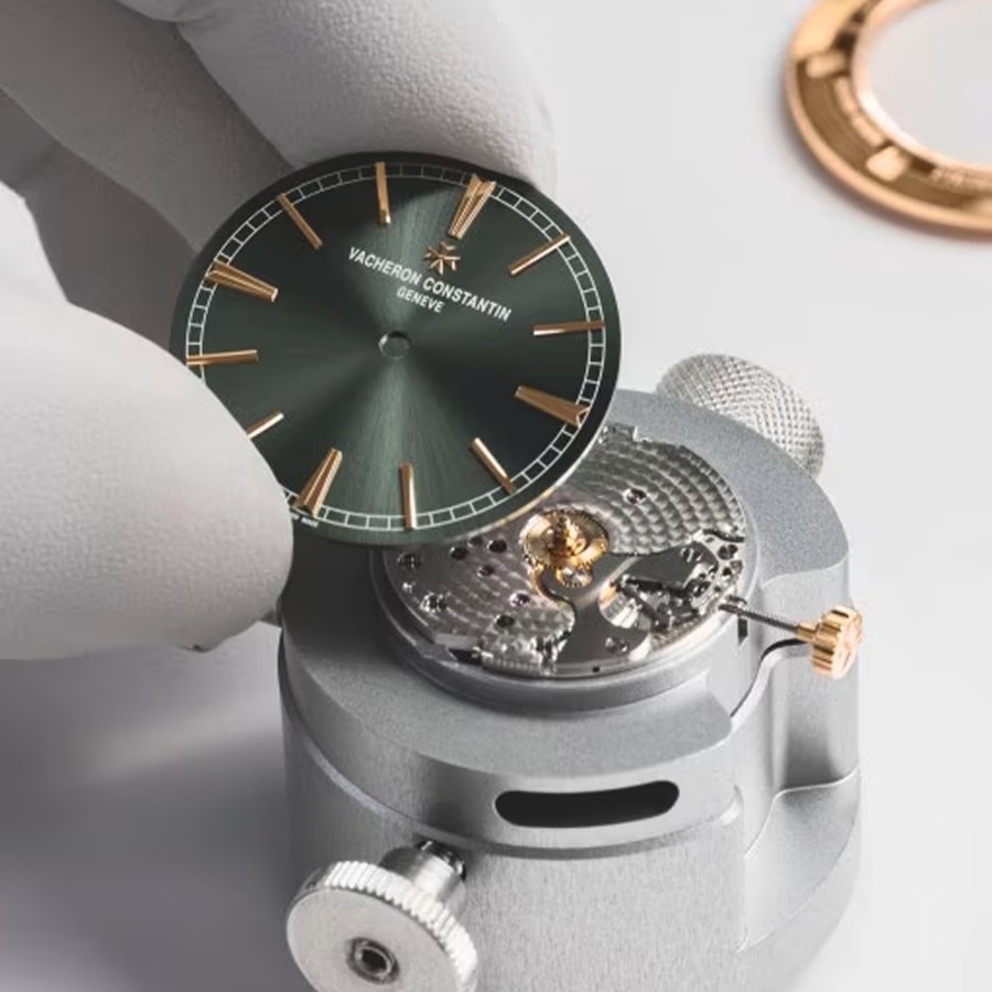 Watchmaker assembling a movement while holding a green dial with gloved hands.