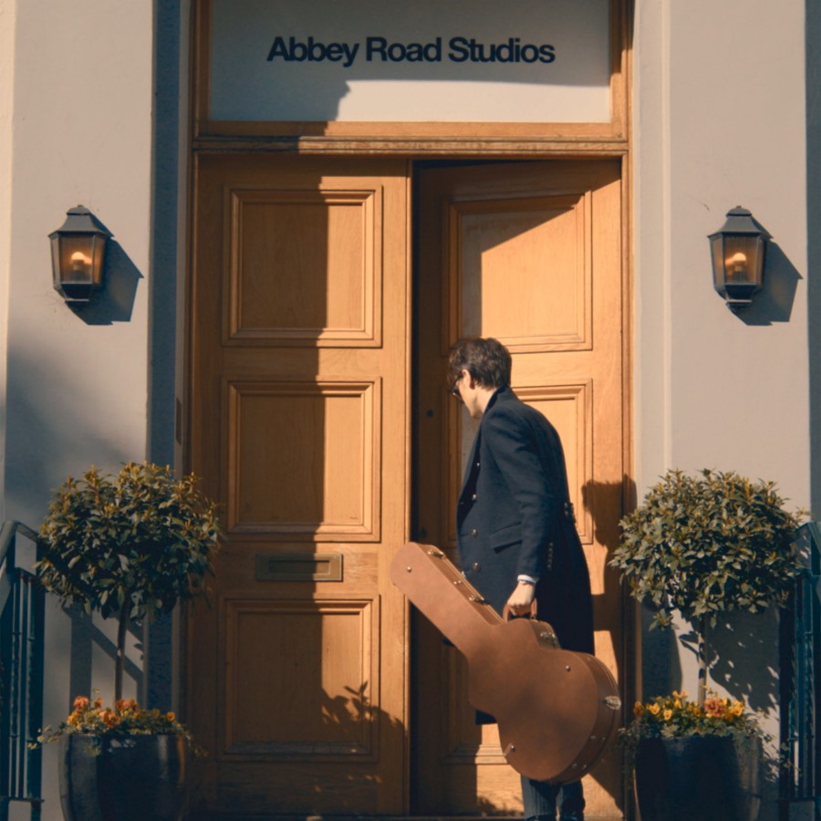 A man carrying a guitar case walking toward the closed doors of Abbey Road Studios.