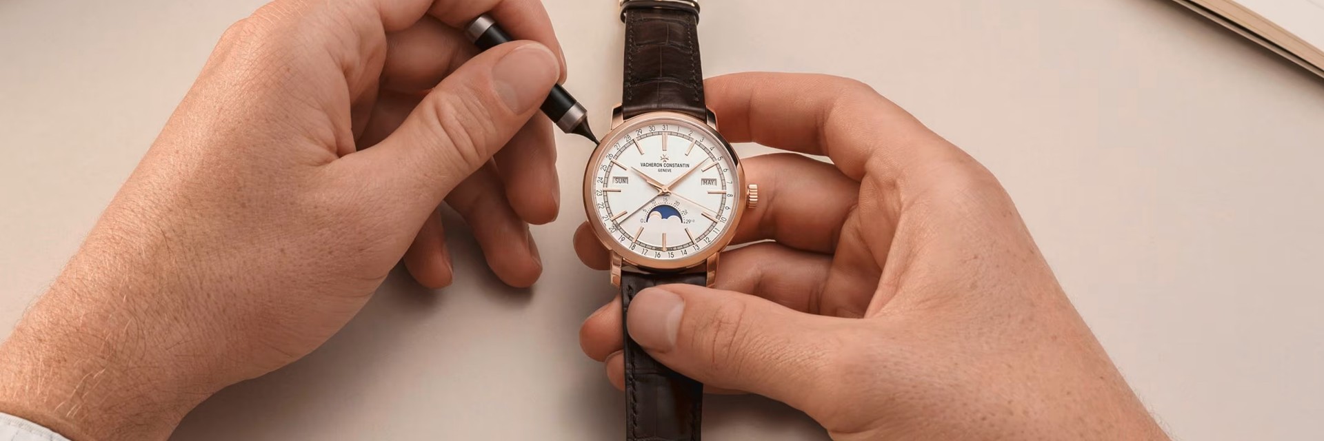 Two human hands are setting up a watch with a leather strap and a white dial with gold hands and frame
