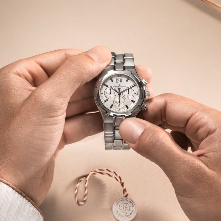 Hands holding a silver Vacheron Constantin watch with a white dial and stainless steel bracelet, placed against a light background with a watch tag beside it.