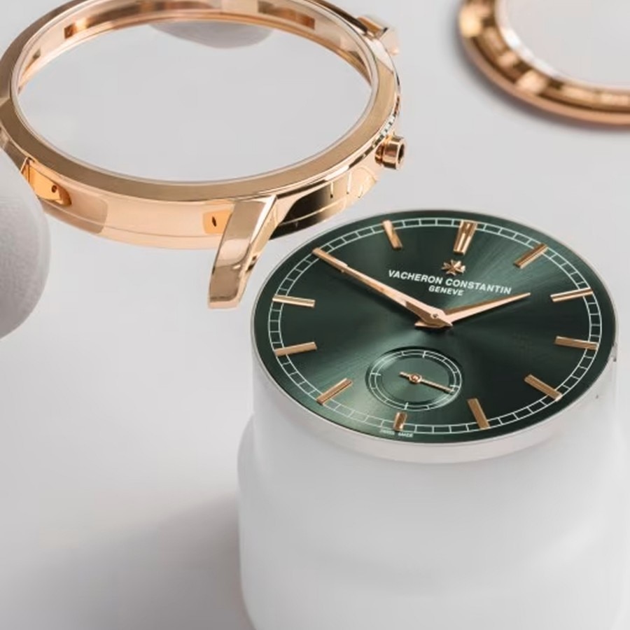 Green watch dial with gold hands displayed alongside disassembled gold case components on a white surface.