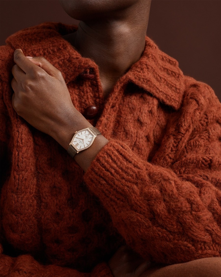 Malte watch with gold case and brown leather strap on wrist in textured orange sweater.